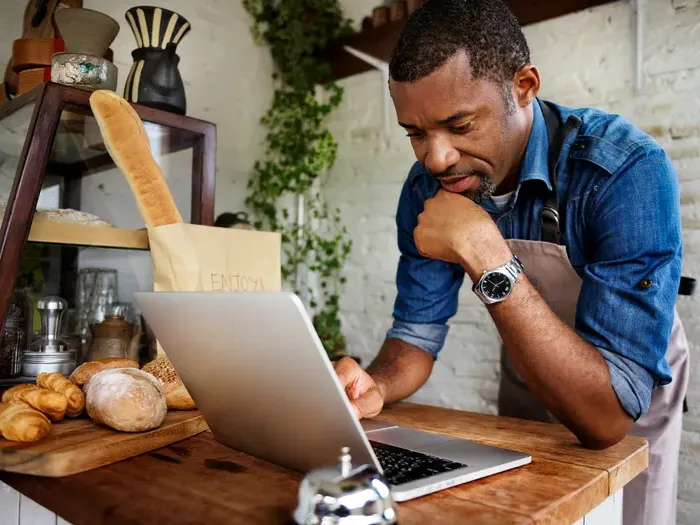 Small bakery owner in an apron works at a laptop beside fresh bread and pastries, managing menus, costs and online customer orders